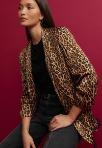 Blazer à imprimé léopard en tissu doux et texturé, à devant ouvert et manches longues, associé à un haut noir et un jean foncé.