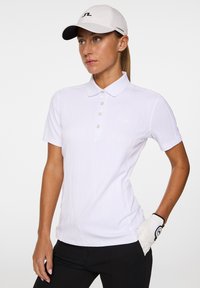 Witte korte mouwen polo met subtiele verticale strepen, knoopkraag en logo-detail. Draag het met zwarte broek en een witte pet.