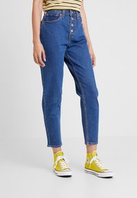 Modré džíny s vysokým pasem a rovnými nohavicemi, s pěti knoflíky a oranžovým šitím. Kombinované s žlutými teniskami Converse.