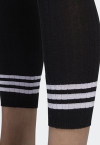 Černé sportovní legíny s ribbovanou texturou a třemi bílými horizontálními pruhy na lemu. Design je těsně přiléhavý a dosahuje ke kolenům.