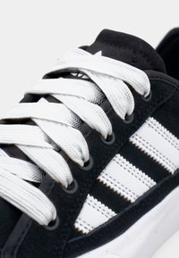 Black suede sneaker with white fabric laces and three white stripes. Features grey grommets, textured accents, and a rubber sole.