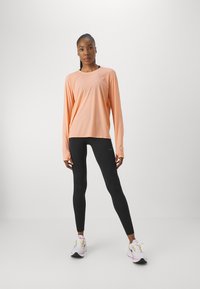 Langärmeliges, hellpfirsichfarbenes Sportoberteil mit lockerer Passform, kombiniert mit schwarzen Leggings und weißen Sneakers mit gelben und rosa Akzenten.