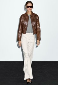 Veste en cuir marron, haut gris, pantalon large blanc, sandales à plateforme noires. Modèle se tient devant un fond uni, portant des lunettes de soleil.