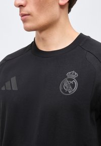 Άνδρας που φοράει μαύρο αθλητικό πουκάμισο με στρογγυλή λαιμόκοψη, με σκουρόγκρι έμβλημα του συλλόγου Ρεάλ Μαδρίτης και λογότυπο Adidas στο στήθος.