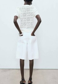 White knitted top with openwork pattern and short sleeves paired with white knee-length shorts. Black sandals and thin black tights are visible.