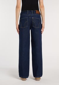 Persona che indossa jeans larghi blu scuro e un top nero, mostrata di spalle con scarpe beige su uno sfondo semplice.