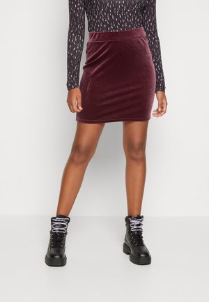 Burgunderroter Minirock aus Samt mit figurbetonter Silhouette. Kombiniert mit schwarzen Schnürstiefeln mit massiver Sohle und grauen Schnürsenkeln. Langärmliges, gemustertes Oberteil.