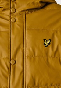 Hnědá prošívaná bunda s patentkami a prošívanou texturou. Na levé části hrudníku je černé a žluté logo orla.