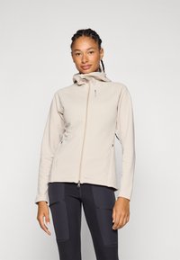 Veste en polaire beige clair avec une fermeture éclair, un col montant et des poches latérales. Texture lisse et design ajusté, associée à des leggings noirs.