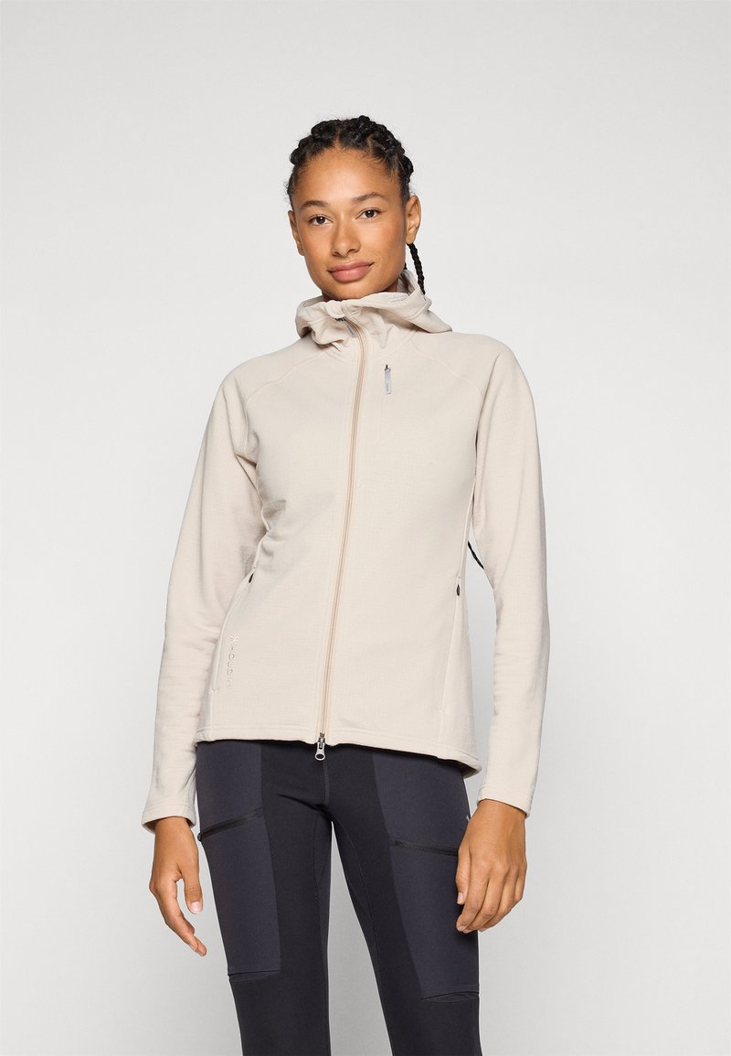 Veste en polaire beige clair avec une fermeture éclair, un col montant et des poches latérales. Texture lisse et design ajusté, associée à des leggings noirs.