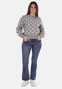 Maglione grigio con pois bianchi, polsini e orlo a costine. Abbinato a jeans blu e sneakers bianche. Modella in piedi con le mani nelle tasche.