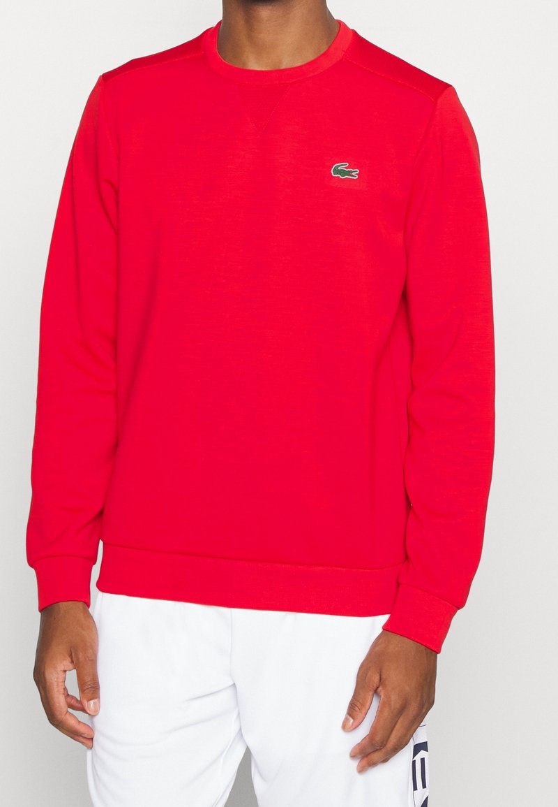 Homme portant un sweat-shirt rouge à manches longues avec un col rond et un petit logo de crocodile vert sur la poitrine, ainsi qu'un pantalon blanc.