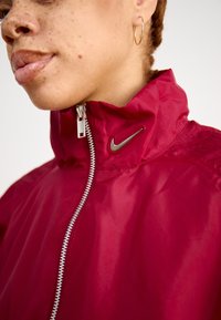 Red windbreaker jacket with a high collar, silver zip, and a small metallic logo on the neck. Features a smooth texture and fitted design.