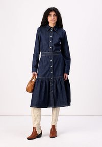 Robe en denim avec un bouton sur le devant, un col et une jupe froncée. Couleur bleu foncé, taille cintrée avec une ceinture. Mannequin portant des bottes crème et tenant un sac marron.