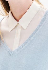 Primo piano di una persona che indossa una camicia bianca con colletto sotto un maglione a V azzurro chiaro, che mostra il collo e la parte superiore del petto.