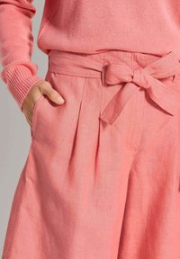 Helle rosa Leinenhose mit hoher Taille, ausgestattet mit Falten, seitlichen Taschen und einem gebundenen Taillendetail. Die Textur wirkt weich und atmungsaktiv.