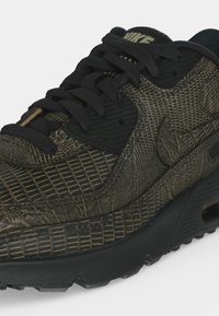 Μαύρο και σκούρο πράσινο αθλητικό παπούτσι Nike με υφή ερπετού, μαύρα κορδόνια και ορατή σόλα με αερόσολα.
