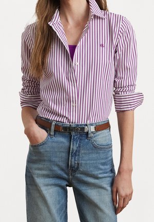 Femme portant une chemise à boutons rayée violet et blanc avec les manches retroussées et un jean bleu, une main dans la poche, sur fond uni.