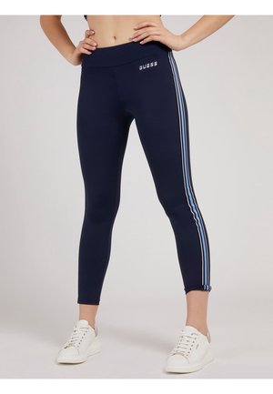 Mujer con leggings azul marino con rayas laterales azules y blancas y zapatillas blancas, de pie con las manos en las caderas.