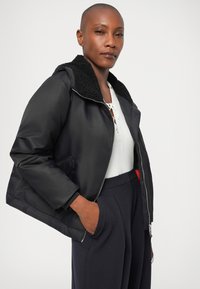 Jachetă neagră căptușită cu un guler texturat negru, închidere cu fermoar, buzunare laterale și un tricou alb cu model în dungi dedesubt. Pantaloni escuți completează ținuta.