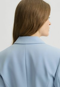 Sacou albastru deschis cu un guler structurat, țesătură moale și detalii de cusătură vizibile, văzut din spate, evidențiind decolteul și umerii.