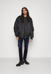 Chaqueta bomber negra con una superficie suave, puños y dobladillo acanalados, combinada con jeans azul oscuro y zapatos negros con cordones.