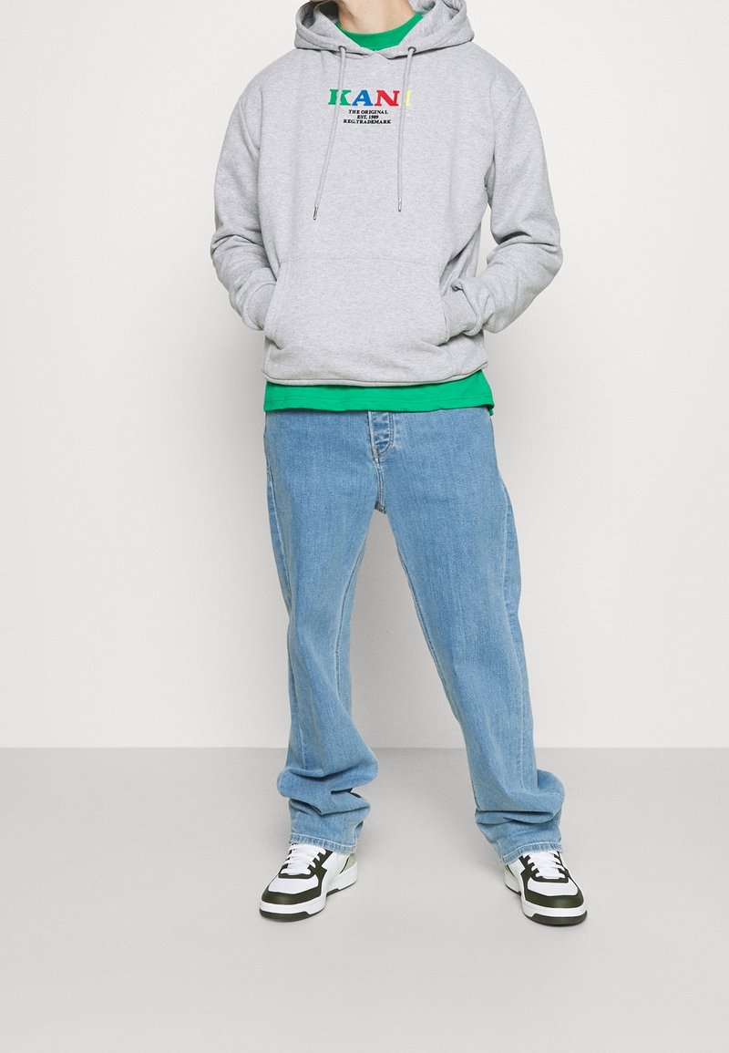 Felpa grigia con grafica frontale, maglietta verde sotto, jeans baggy azzurri, e scarpe nere/bianche con accenti verdi.