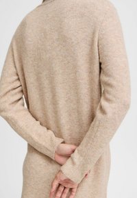 Beige gebreide trui van een zachte, textuur stof. Heeft geribbelde manchetten en een relaxte pasvorm, met een gladde rug en minimale details.