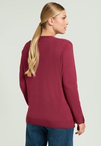 Maglione bordeaux a maniche lunghe, orlo a coste, spacchi laterali e tessuto morbido, abbinato a pantaloni di jeans blu. Vista posteriore mostrata.
