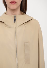 Giacca beige con cappuccio caratterizzata da una zip frontale e un dettaglio a patch rettangolare. Realizzata in tessuto liscio con un design leggero.