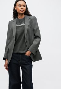 Blazer gris a rayas con un corte ajustado, combinado con una camiseta gris que presenta texto bordado en blanco, y vaqueros de mezclilla oscuros.