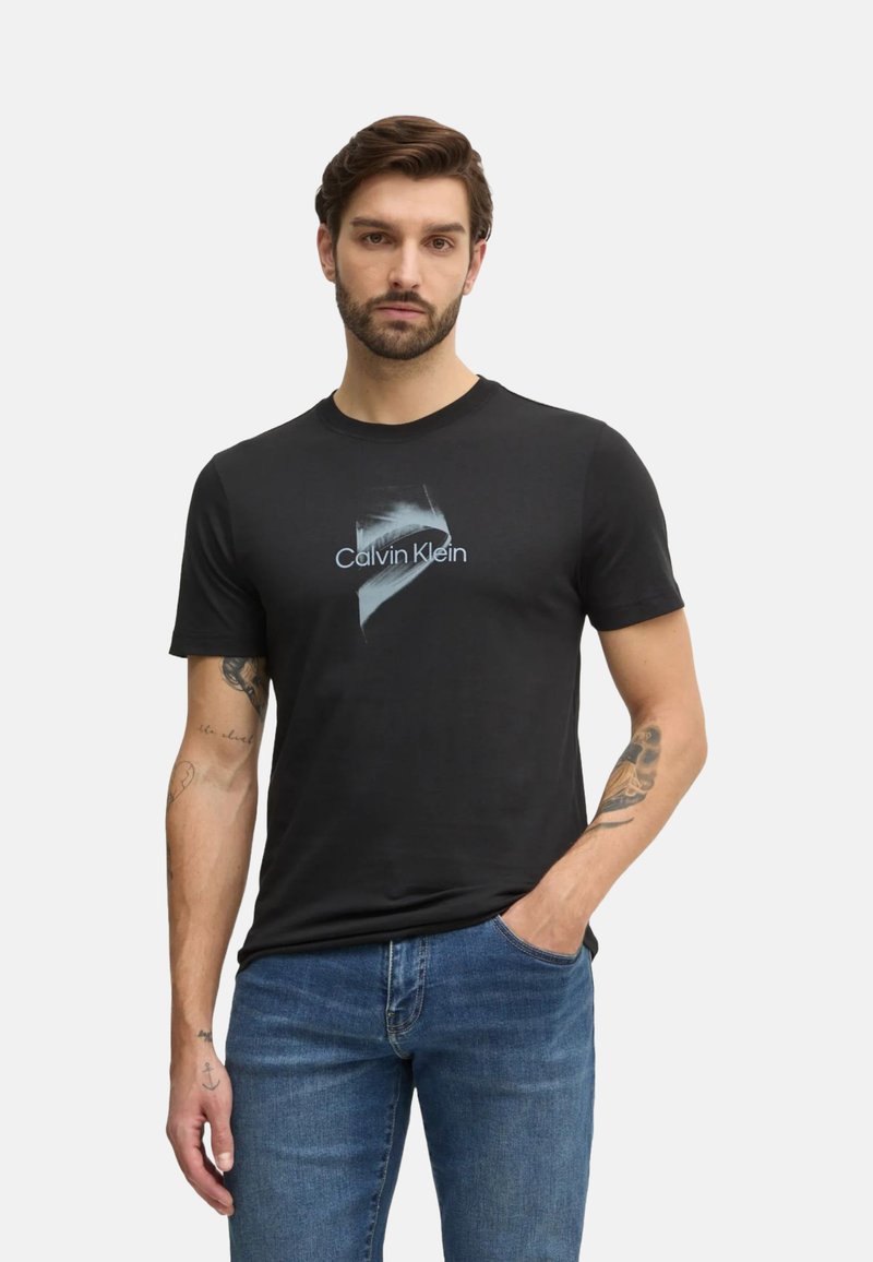 Camiseta negra de algodón con cuello redondo, que presenta un sutil gráfico azul y el texto "Calvin Klein" impreso en blanco. Llevada con jeans azules.