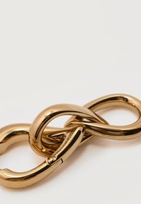 Κοντινό πλάνο διασυνδεδεμένων γυαλιστερών χρυσών συνδέσμων αλυσίδας με κυρτό κούμπωμα χαραγμένο με τη λέξη "Calvin Klein" σε λευκό φόντο.