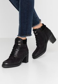 Botines negros hasta el tobillo con un acabado de ante texturizado, que presentan cordones, cremallera lateral y un grueso tacón cuadrado. Llevados con jeans ajustados.