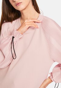 Helles rosa Sweatshirt mit plissierten Ärmeln, geripptem Kragen und elastischen Bündchen. Verfügt über eine Kordel mit schwarzem Metallbeschlag an den Ärmeln.