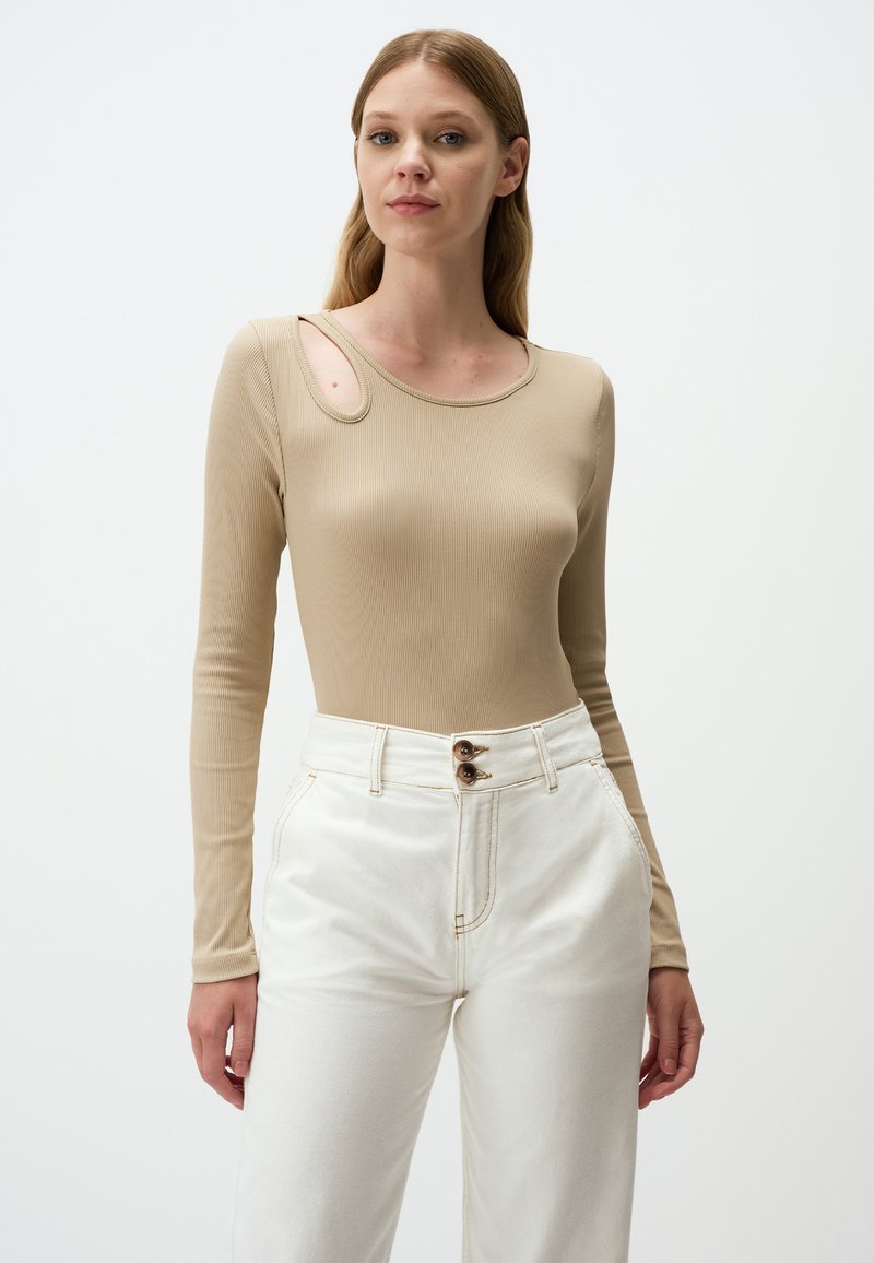 Beige langärmliges, geripptes Oberteil mit einem markanten Ausschnitt an der Schulter, kombiniert mit hochgeschnittenen weißen Hosen mit zwei Knopfverschlüssen.