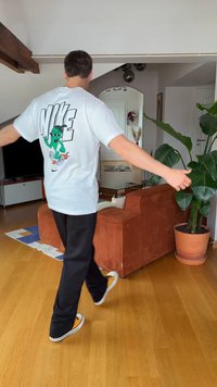 Férfi sétál bent kinyújtott karokkal, fehér Nike pólót visel rajzfilm mintával, fekete nadrágot és sárga tornacipőt egy barna kanapé és növény közelében.