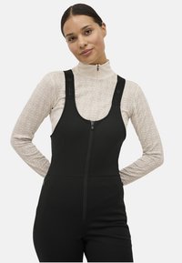 Kvinde iført en beige mønstret turtleneck under sorte zip-up overalls med brede skulderstropper, stående med hænderne bag ryggen.