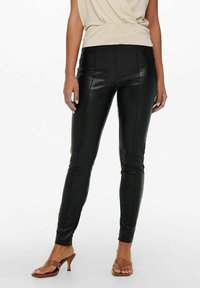 Leggings en simili cuir noir avec une coupe skinny, dotés de coutures verticales et d'une texture lisse. Associés à un haut beige et des sandales marron.