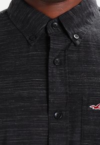 Camicia grigia scura con bottoni, caratterizzata da sottili righe orizzontali e un piccolo logo ricamato rosso sulla tasca del petto con il colletto visibile.