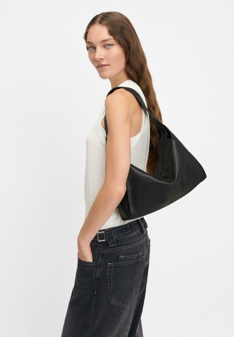 Czarna skórzana torebka na ramię o unikalnym trójkątnym kształcie, z teksturowaną powierzchnią i pojedynczym paskiem, w połączeniu z szarymi dżinsami o wysokim stanie.