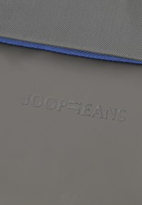 Grauer Stoff mit einer strukturierten Oberfläche, versehen mit blauen Nähten entlang des Randes. Das geprägte Logo trägt die Aufschrift "JOOP®JEANS."
