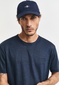Uomo con barba chiara e tatuaggi che indossa una maglietta blu navy e un berretto blu navy con un emblema rosso e bianco, rivolto in avanti su uno sfondo semplice.