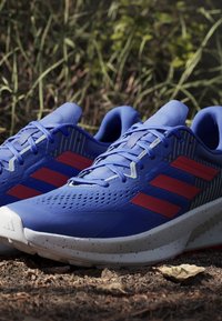 Blue athletic shoes with red accents, featuring a textured upper, perforations for breathability, and a white speckled sole.