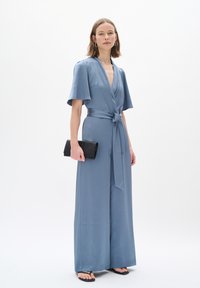 Blaue Wickel-Overall mit weiten Ärmeln und gebundenem Taillenbund. Glatter Stoff, bodenlanges Design, getragen mit einer schwarzen Clutch und Sandalen.