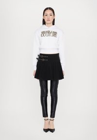 Felpa bianca corta con logo metallico, gonna plissettata nera a vita alta, leggings neri lucidi e décolleté neri a punta.