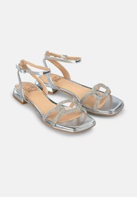 Zilveren sandalen met een glanzende metalen afwerking, een enkelbandje en een decoratieve kristallen sluiting op de teband; vierkant gevormd teendesign.