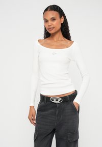 Hosszú, göndör hajú nő fehér hosszú ujjú felsőt és fekete, ezüst csattal díszített fekete övvel ellátott cargo nadrágot visel.