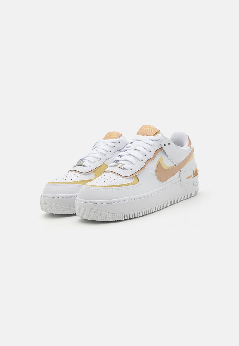 Zapatillas de cuero blanco con acentos en naranja claro, punta redondeada, cierre con cordones y suela texturizada que presenta un detalle de marca prominente.