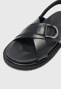 Black leather sandal featuring crisscross straps, a metallic buckle accent, and a textured rubber sole. Minimalist design with smooth finish.